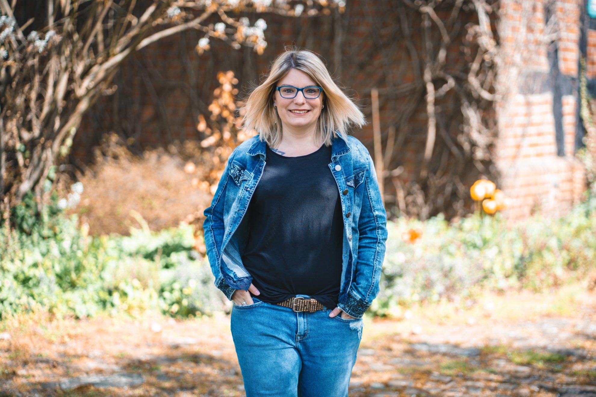 Carolin trägt ein schwarzes Shirt und darüber eine Jeansjacke. Carolin steht vor einem alten Fachwerkhaus.