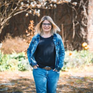 Carolin trägt ein schwarzes Shirt und darüber eine Jeansjacke. Carolin steht vor einem alten Fachwerkhaus.