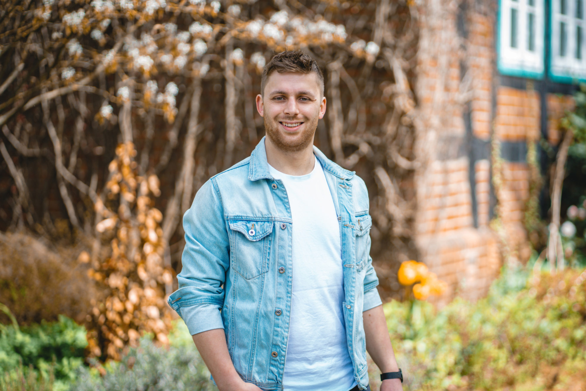Lars Fleddermann trägt ein weißes Shirt und darüber eine Jeansjacke. Er steht vor einem alten Fachwerkhaus.
