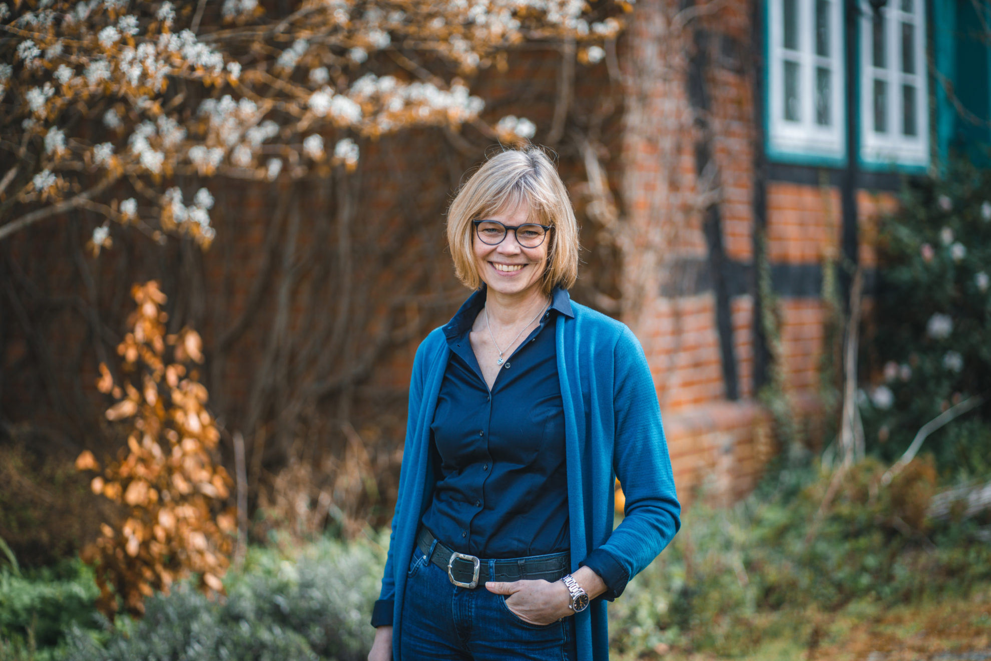 Ingrid Hüdepohl steht vor einen alten Fachwerkhaus. Sie trägt eine dunkelblaue Bluse und darüber eine hellblaue Jacke.