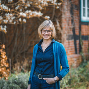 Ingrid Hüdepohl steht vor einen alten Fachwerkhaus. Sie trägt eine dunkelblaue Bluse und darüber eine hellblaue Jacke.